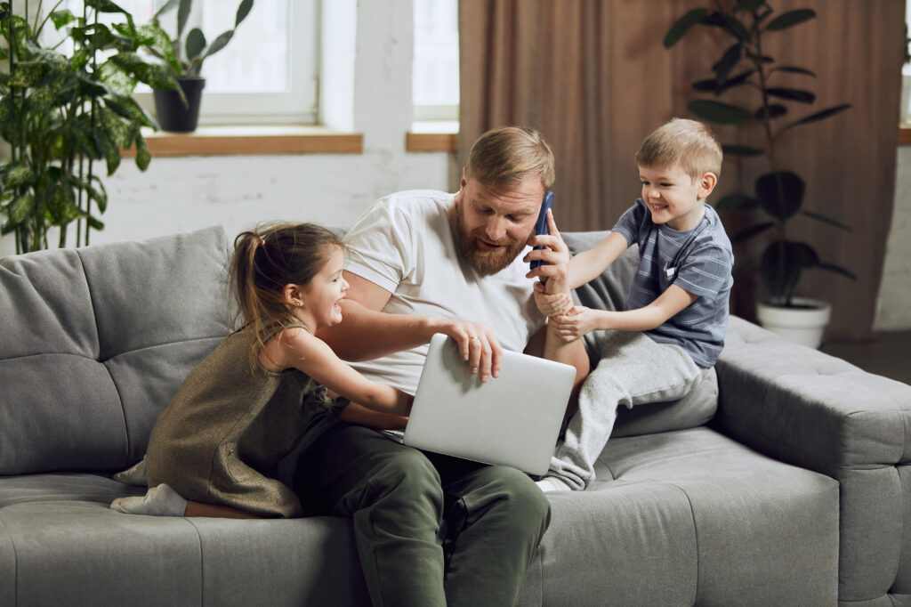 Man, father sitting on couch, talking on phone and working on laptop in living room. Kids bothering and playing with him