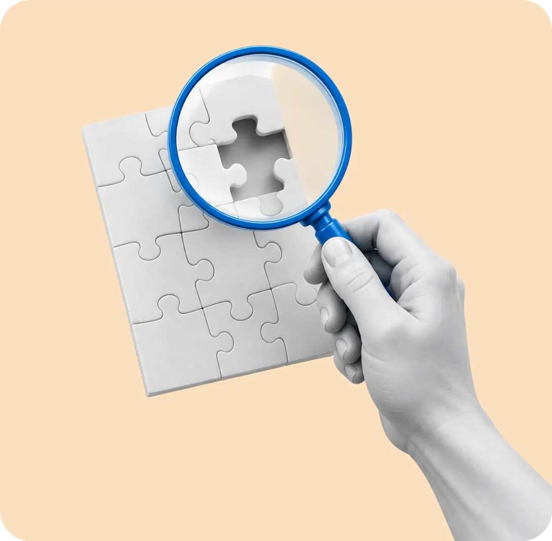 A hand holding a magnifying glass over a jigsaw puzzle with a missing piece